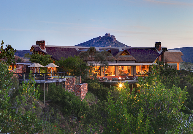 View of the Botlierskop Tented Lodge Restaurant, a charming structure blending natural stone and thatch roofing, surrounded by lush greenery. The lodge features expansive glass windows offering warm, inviting interiors and a large deck area with outdoor seating, overlooking a serene wilderness backdrop. The lodge’s elevated position provides stunning views of the surrounding landscape, with a majestic hill visible in the background